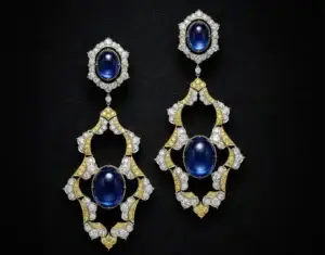 The earrings "The Sapphier Ascend" on display. They have the shape of golde feathers with no filling. In the center there is one big ova sapphire and it has an upper part that can be detached. This part is a hexagon encrsuted with gold and diamonds with another oval sapphire in the center