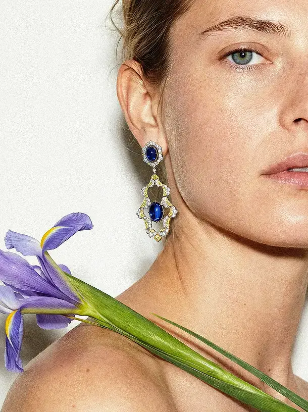 A model posing with the left side of her face cut out, a purple and green flower over her shoulder and wearing one of the Sapphire Ascend earrings